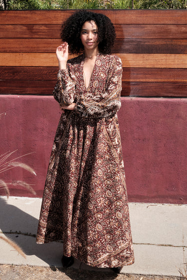 A woman standing by a red wall, outside wearing a long sleeve maxi caftan cut in a raisin floral tapestry.