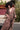 A woman standing by a red wall, outside wearing a long sleeve maxi caftan cut in a raisin floral tapestry, belted.