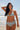 A woman on a beach wearing a white ground with lavender, blue and green floral tapestry bikini bottom with a high rise and matching bikini top.