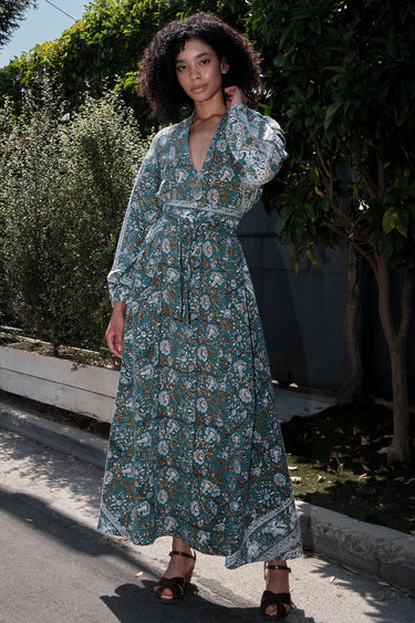 Woman wearing a long tarragon colored floral tapestry caftan, belted, standing outdoors with greenery in the background.