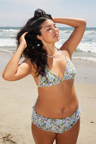 A woman at the beach wearing a white ground, lavender and blue floral tapestry triangle bikini top and matching bikini bottom.