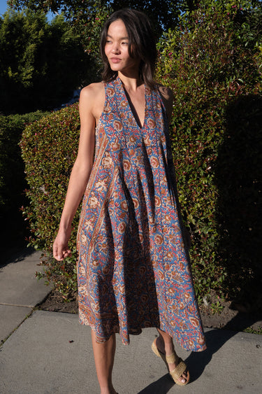 A woman standing in front of greenery outside wearing a sleeves midi caftan cut in a blue floral tapestry.