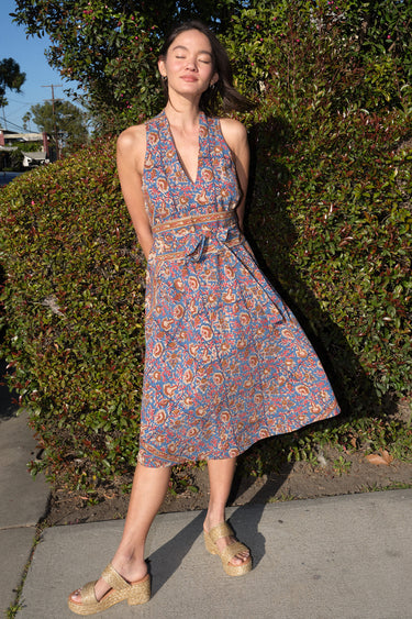 A woman standing in front of greenery outside wearing a sleeves midi caftan cut in a blue floral tapestry, belted.