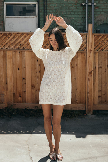 A woman standing outside by a brown wood fence wearing a lace mini dress with gold sandals.