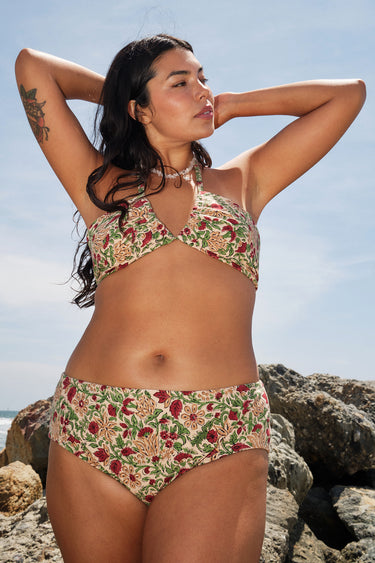 A woman on a beach by rocks wearing a beige ground with red, brown and green floral tapestry bikini bottom with a high rise and matching bikini top.