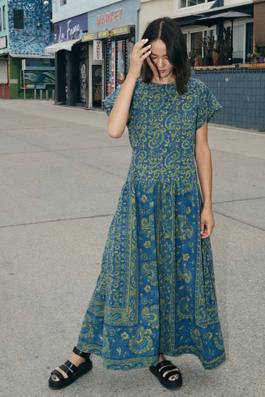 A woman at a beach boardwalk wearing a dark blue paisley tapestry maxi dress with pockets in the gathered skirt.
