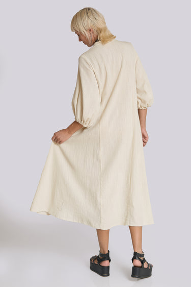 A woman standing in a white wall photo studio wearing a 3/4 sleeve midi alabaster off white caftan. Back View
