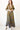 A woman standing in a studio with white walls wearing a maxi caftan cut in an olive green tree of life handloom tapestry.