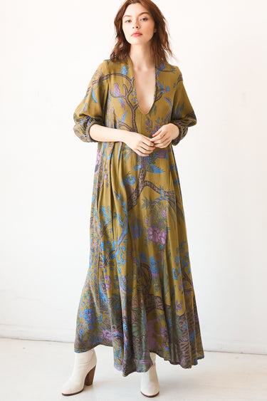 A woman standing in a studio with white walls wearing a maxi caftan cut in an olive green tree of life handloom tapestry.