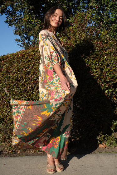 A woman standing outside by greenery wearing a maxi caftan cut in a tree of life handloom tapestry.