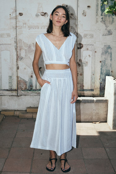 A woman standing outside by a mural wearing a white gauze with black stripe skirt with side slits and a drop waist, paired with a matching crop top.