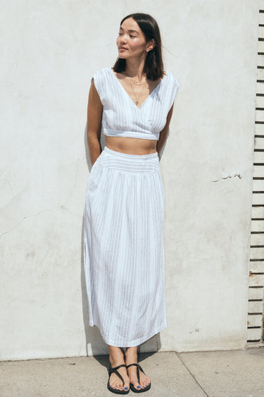 A woman leaning on a white wall, outside wearing a white with black stripe gauze crop top with matching skirt.