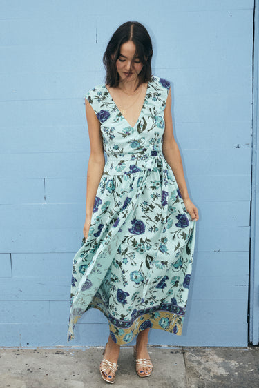 A woman standing by a light blue wall wearing a wrap dress cut in an aqua ground floral tapestry.