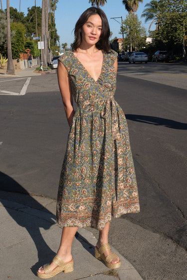 A woman standing on the sidewalk wearing a green floral tapestry midi dress with an elastic neckline and pockets.