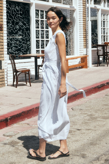 A woman standing in a street with shops wearing a long white with black stripe gauze sleeveless wrap dress, side view.