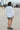 A woman on a beach side walk wearing a long sleeved white with black stripe gauze shirt jacket paired with a gray short, back view.