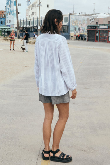 A woman on a beach side walk wearing a long sleeved white with black stripe gauze shirt jacket paired with a gray short, back view.