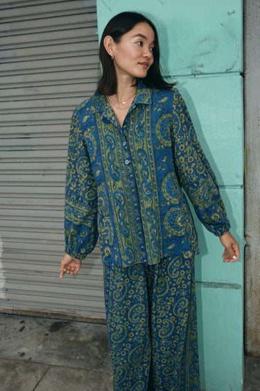 A woman standing outside by a wall wearing a shirt jacket cut from a dark blue paisley tapestry, with a matching trouser.