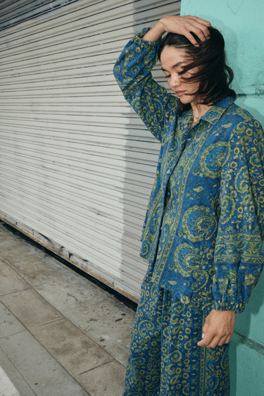 A woman standing outside by a wall wearing a shirt jacket cut from a dark blue paisley tapestry, with a matching trouser.
