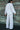 A woman outside by a verdigris wall wearing a long sleeved white with black stripe gauze shirt jacket with a matching trouser, back view.