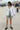 A woman on a beach side walk wearing a long sleeved white with black stripe gauze shirt jacket paired with a gray short and a white bra top.