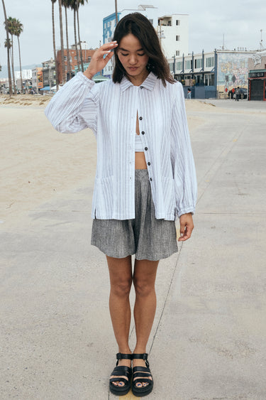 A woman on a beach side walk wearing a long sleeved white with black stripe gauze shirt jacket paired with a gray short and a white bra top.