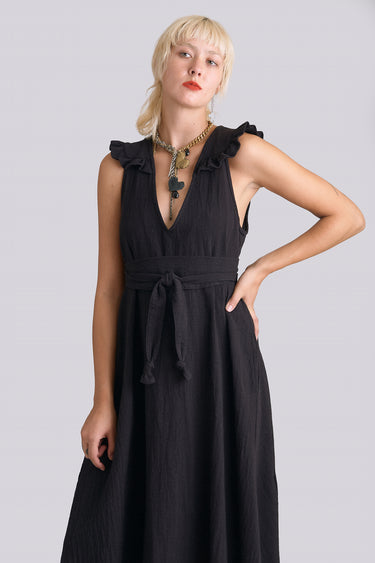 A woman standing in a photo studio with white walls wearing a black circular flared sleeveless midi dress with ruffles around the yoke, with an obi belt.