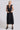 A woman standing in a photo studio with white walls wearing a black circular flared sleeveless midi dress with ruffles around the yoke, with an obi belt.