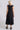 A woman standing in a photo studio with white walls wearing a black circular flared sleeveless midi dress with ruffles around the yoke, with an obi belt. Back View
