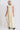 A woman standing in a photo studio with white walls wearing an off white alabaster circular flared sleeveless maxi dress with ruffles around the yoke, back view.