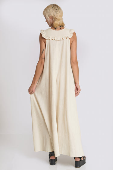 A woman standing in a photo studio with white walls wearing an off white alabaster circular flared sleeveless maxi dress with ruffles around the yoke, back view.