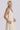 A woman standing in a photo studio with white walls wearing an off white alabaster circular flared sleeveless maxi dress with ruffles around the yoke, with an obi belt.
