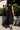 A woman standing outside in front of a lime tree wearing a black circular flared sleeveless midi dress with ruffles around the yoke. Back View