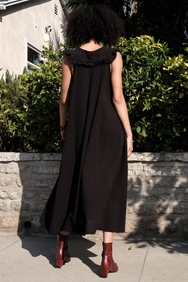 A woman standing outside in front of a lime tree wearing a black circular flared sleeveless midi dress with ruffles around the yoke. Back View