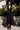 A woman standing outside in front of a lime tree wearing a black circular flared sleeveless midi dress with ruffles around the yoke.