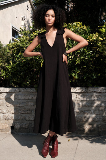 A woman standing outside in front of a lime tree wearing a black circular flared sleeveless midi dress with ruffles around the yoke.