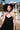 A model wearing a black circular maxi slip dress standing outside near a white picket fence with a house in the background. Neckline detail