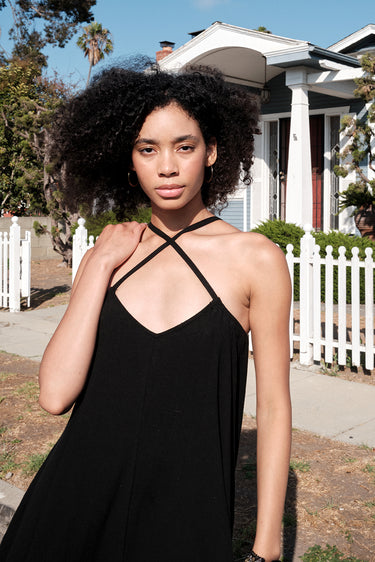 A model wearing a black circular maxi slip dress standing outside near a white picket fence with a house in the background. Neckline detail