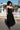 A model wearing a black circular maxi slip dress standing outside near a white picket fence with a house in the background.