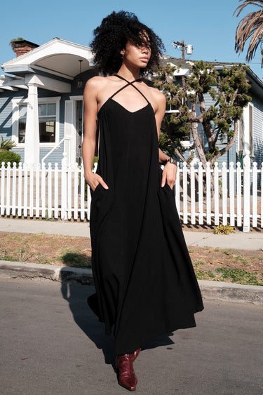 A model wearing a black circular maxi slip dress standing outside near a white picket fence with a house in the background.
