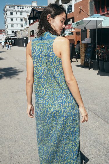 A woman on a beach boardwalk wearing a sheath dress made from a dark blue paisley tapestry. Back View