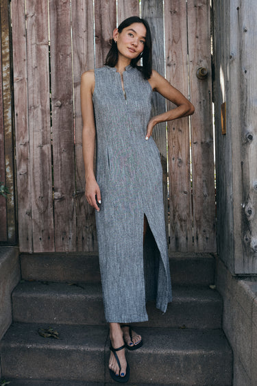 A model wearing a gray maxi shift dress with a front zipper and pockets, standing near a wooden fence.