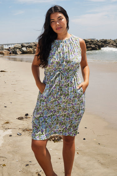 A woman standing on a beach wearing a knee-length floral dress with broomstick pleats, featuring an elastic neckline and a slit back. The dress is a white ground tapestry with blue and lavenders flowers.