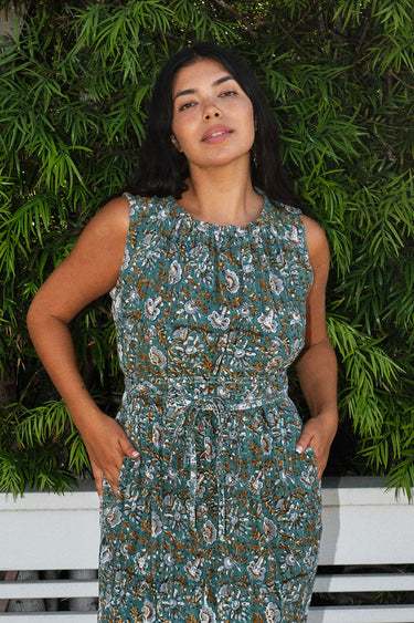 A woman standing outdoors with greenery wearing a green and brown floral patterned tapestry maxi sheath with broomstick pleats, belted.