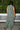 A woman standing outdoors with greenery wearing a green and brown floral patterned tapestry maxi sheath with broomstick pleats, back view.