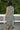 A woman standing outdoors with greenery wearing a green and brown floral patterned tapestry maxi sheath with broomstick pleats, belted, back view.