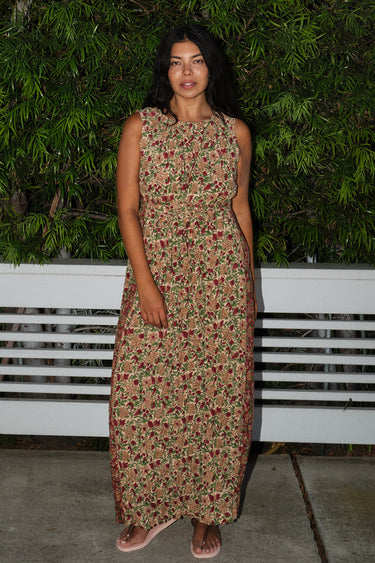 A woman standing by greenery wearing a maxi floral dress with broomstick pleats, featuring an elastic neckline and a slit back. The dress is a beige ground tapestry with red and brown flowers.