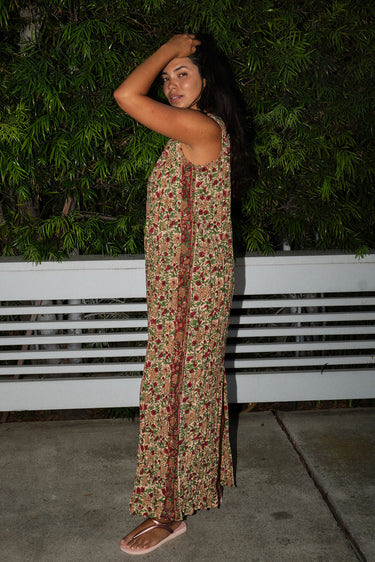 A woman standing by greenery wearing a maxi floral dress with broomstick pleats, featuring an elastic neckline and a slit back. The dress is a beige ground tapestry with red and brown flowers. Side view