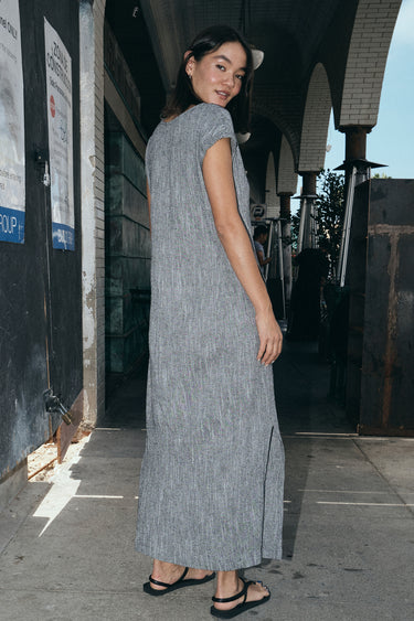 Woman standing on the sidewalk, outside wearing a salt and pepper yarn dye long sheath dress with a cap sleeve. Back View.