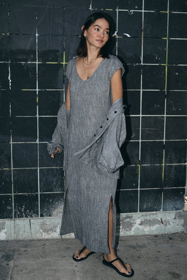 Woman standing by a black tile wall outside wearing a salt and pepper yarn dye long sheath dress with a cap sleeve. She has a matching jacket draped on her arms.